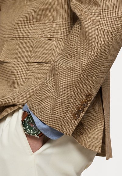 Bruin geruite blazer met een gestructureerde afwerking, voorzien van drie bruine knopen op de mouw. Een lichtblauwe shirtmanchet steekt eronderuit. Een groene horloge met een bruine band is zichtbaar.