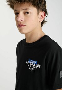 T-shirt en coton noir à manches courtes, avec un imprimé graphique blanc et bleu affichant "ART GALLERY NEW YORK" à l'avant.