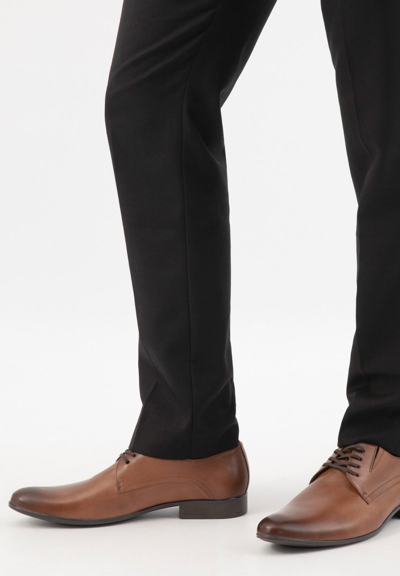 Bruine leren nette schoenen gedragen met zwarte, op maat gemaakte broek bij een persoon die tegen een witte achtergrond staat.