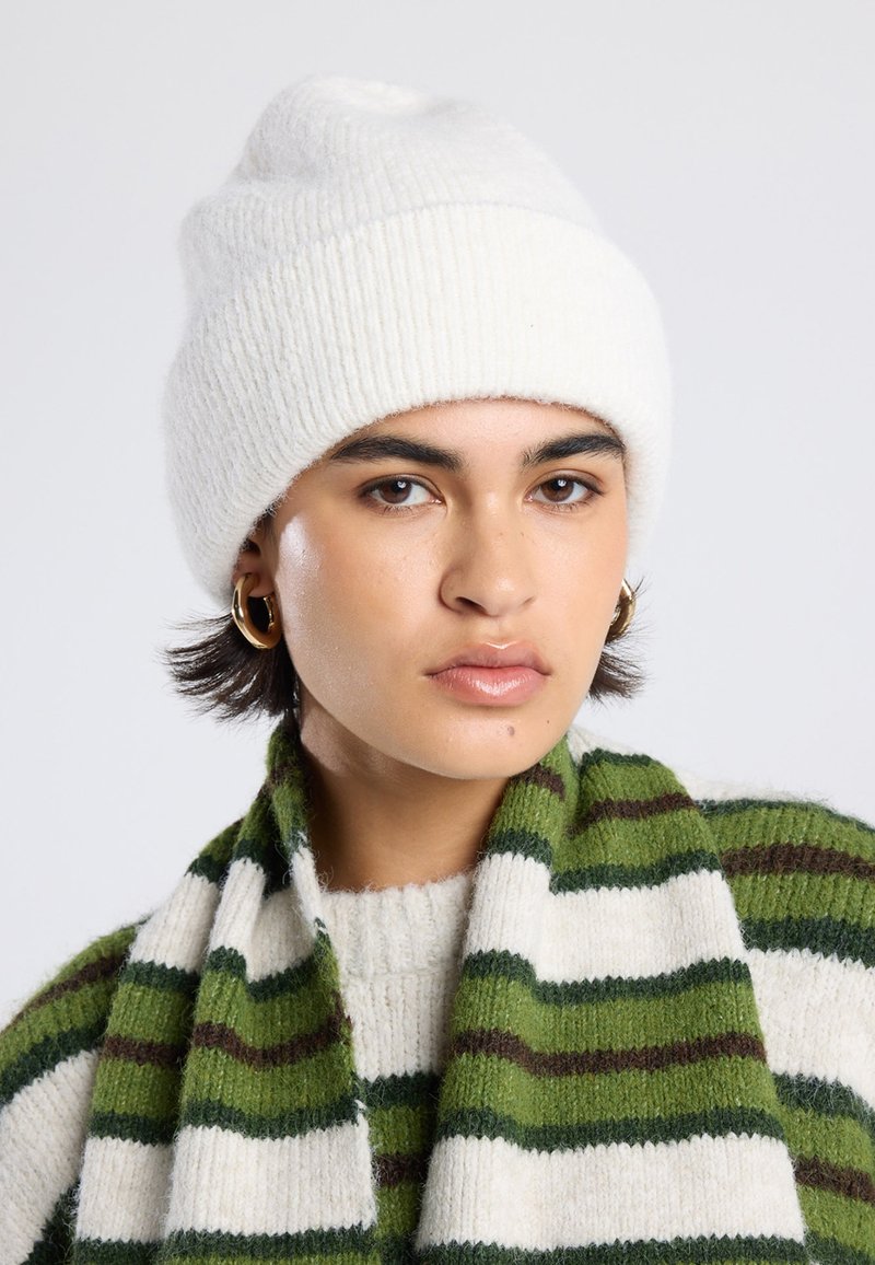 White wool beanie with ribbed texture, paired with a green and cream striped scarf. Model has short hair and gold hoop earrings.
