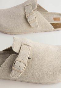 Zuecos de fieltro gris claro con una correa que presenta una hebilla de metal rectangular y una suela texturizada. Diseño tipo pantufla con la parte trasera abierta.