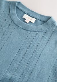 Pull en tricot bleu clair avec un col rond, présentant des détails côtelés verticaux et une étiquette en tissu indiquant la taille M.
