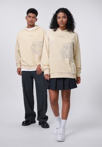 Felpe crema con grafiche sul davanti, abbinate a pantaloni neri a righe e una gonna plissettata, sneakers bianche e calzini alla caviglia.