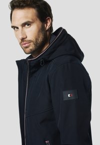 Marineblaue Kapuzenjacke aus weichem Stoff, mit Reißverschluss vorne, kontrastierenden roten Akzenten und einem Logo-Patch am linken Ärmel.