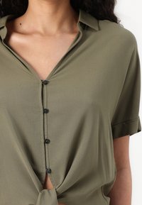 Olive green short-sleeved shirt with a collar, featuring a button placket, a knotted front, and a smooth, lightweight fabric texture.