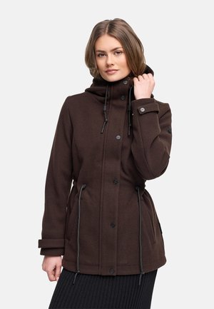 Naine, kes kannab pruuni nööpidega mantlit, millel on kõrge krae ja nöörid, hoiab kraest ühe käega kinni, seistes tavalise tausta ees.