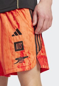 Orange og røde tie-dye shorts med sorte striber, der har Adidas-logo, "NTS" label og kanon grafik. Lavet af letvægtsstof.