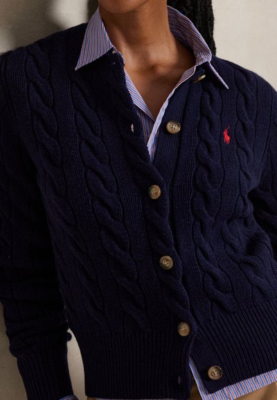 Persona que lleva un cárdigan de punto trenzado azul marino con botones marrones sobre una camisa con rayas azules y rojas y cuello.