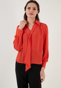 Red, lightweight blouse with long sleeves, a tie neckline, and buttoned cuffs. Smooth texture and slight draping at shoulders.
