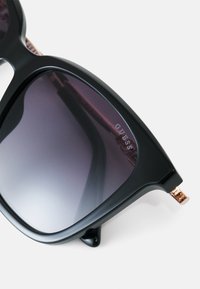 Occhiali da sole in plastica nera con lenti sfumate che passano da un viola scuro a un viola chiaro. Presentano un accento metallico sulla tempia con il logo del brand.