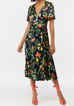 Woman wearing navy wrap dress with colorful floral and fruit print, puffed short sleeves, and coral ankle-tie high-heeled sandals.