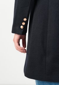 Main à côté d'une manche de manteau bleu marine avec trois boutons dorés, porté sur un jean bleu, présenté sur un fond clair uni.
