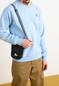 Ljusblå sweatshirt med ribbade muddar, ihop med khakifärgade byxor. En liten svart crossbody-väska med en logotyp och randig rem bärs.