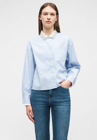 Mujer con camisa azul claro de rayas finas y cuello blanco adornado, y jeans azules, de pie con una mano en el bolsillo contra un fondo simple.