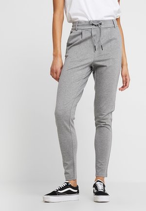 Vrouw die grijze slim-fit joggerbroek met koordje in de taille en zwart-witte sneakers draagt, staand tegen een effen achtergrond.