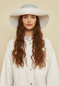 Chapéu branco de aba larga com parte superior riscada. Usado com uma camisa branca oversized, com detalhes dos botões visíveis. Cabelo castanho ondulado.