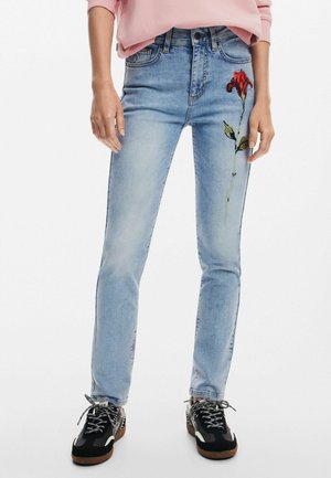 Jean azul de denim con forma ajustada, que presenta una rosa bordada roja en el muslo izquierdo y se lleva con zapatillas negras.