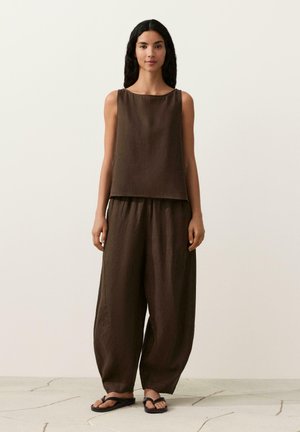 Vrouw met lang zwart haar, gekleed in een los bruin mouwloos topje, wijde broekspijpen en zwarte sandalen, staande tegen een effen beige achtergrond.