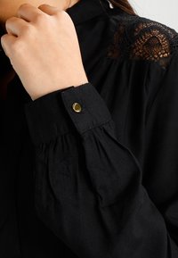 Blusa nera con dettagli in pizzo testurizzato sulle spalle, maniche arricciate e chiusura con bottone dorato ai polsi. Tessuto morbido, design elegante.