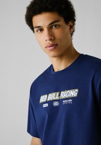 Młody mężczyzna z kręconymi, ciemnymi włosami, ubrany w granatowy t-shirt Red Bull Racing, patrzący w przód na jasnoszarym tle.