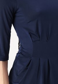 Navy blue dress featuring smooth fabric, three-quarter sleeves, gathered side details, and structured pleats at the waistline.