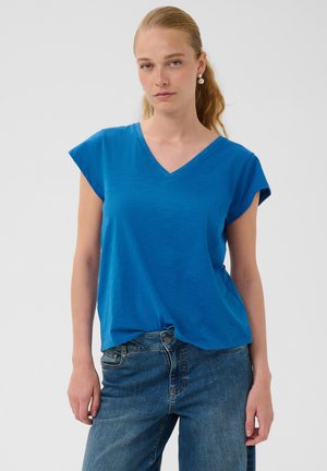 Femme à la peau claire et aux longs cheveux blonds portant des boucles d'oreilles en perles, un haut bleu à col en V à manches courtes, et un jean bleu, devant un fond uni.
