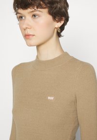 Beige gerippter Strick-Longsleeve mit hohem Ausschnitt und einem kleinen, rosa Levi's-Logoetikett auf der Brust. Glatte Textur, taillierte Passform.