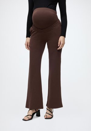 Personne enceinte portant un pantalon de maternité marron avec une ceinture élastique et un haut noir à manches longues, debout dans des sandales noires à talons.