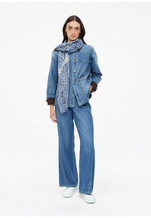 Frau trägt blaue Jeansjacke und weite Jeans mit weißen Schuhen und blau gemustertem Schal und steht vor weißem Hintergrund.