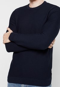 Marinblå, stickad tröja med lång ärm, rund hals, ribbade muddar och nederkant. Har ett strukturerat mönster genom hela tyget.