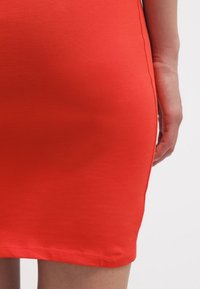 Gros plan sur le bas du torse et la main d'une personne portant une jupe rouge ajustée sur un fond blanc uni.