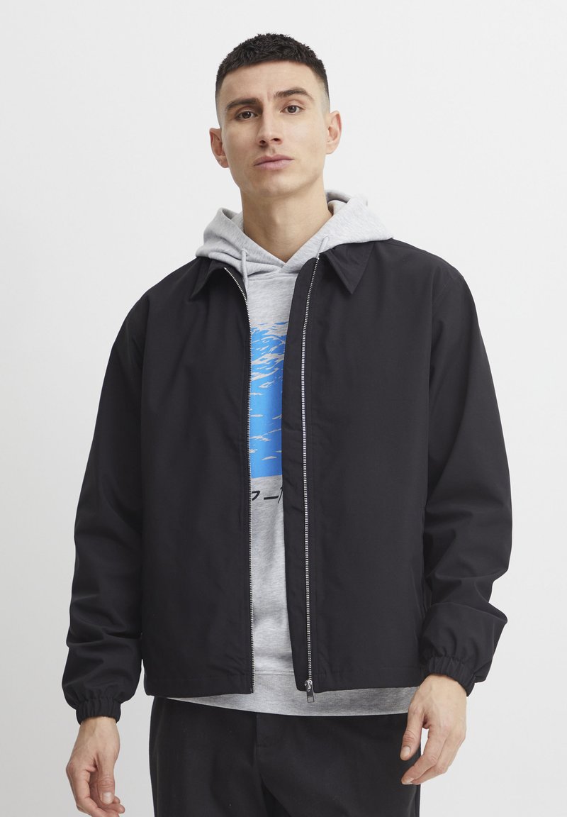 Schwarze leichte Jacke mit Kragen, ausgestattet mit einem Frontreißverschluss und elastischen Bündchen, getragen über einem grauen Hoodie mit blauem Grafikdesign.