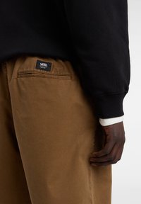 Bruine katoenen broek met een voorzak en een label met de tekst "VANS." Het model draagt een zwart sweatshirt en een wit gemoffeld shirt eronder.