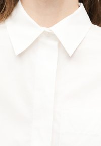 Κοντινό πλάνο ενός ατόμου που φοράει λευκό πουκάμισο με γιακά, με το πάνω κουμπί κουμπωμένο και εμφανή καστανά μαλλιά στις πλευρές.