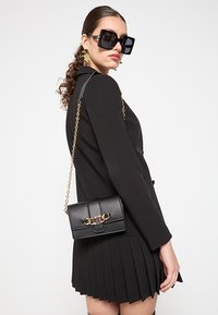 Borsa a tracolla in pelle nera con finitura testurizzata, dettaglio a catena dorata e chiusura a pattina. Indossata con un completo nero su misura.