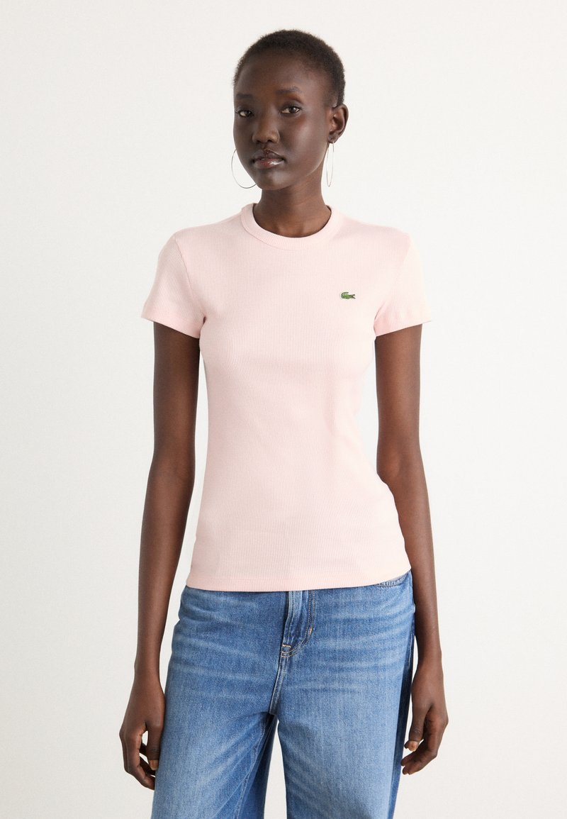 T-shirt côtelé rose clair avec un col rond et des manches courtes, arborant un petit logo de crocodile vert Lacoste sur la poitrine.