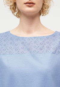 Γυναίκα με μπούκλες ξανθά μαλλιά που φοράει ανοιχτό μπλε μπλουζάκι με φλοράλ δαντέλα στο ντεκολτέ και λαιμόκοψη σκάφος.