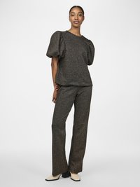 Mulher vestindo uma blusa de mangas bufantes texturizadas escuras e calças largas combinando, com sapatos de bico quadrado em creme, em pé contra um fundo branco.