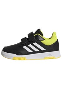 Zapatilla deportiva negra con rayas blancas y acentos amarillo brillante. Presenta una parte superior texturizada y una suela exterior de goma blanca. Tirantes de velcro.