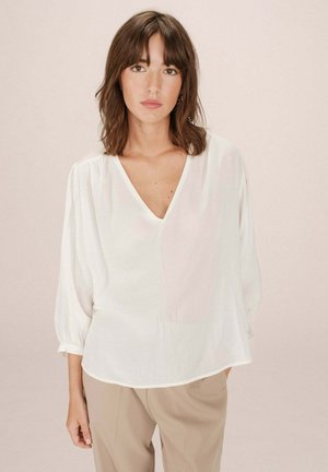 Blouse blanche ample avec un décolleté en V profond et des manches bouffantes. Texture douce et tissu léger. Associée à un pantalon beige ajusté.