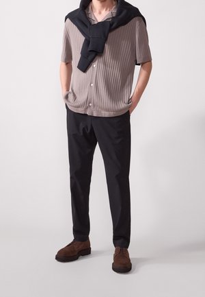 Chemise beige côtelée à manches courtes avec des boutons, associée à un pantalon noir et des chaussures en daim marron. Pull noir posé sur les épaules.