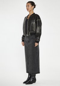 Giacca bomber in pelle nera con cuciture a contrasto, polsini a coste e tasche laterali, abbinata a una gonna in denim scuro e stivali alla caviglia.