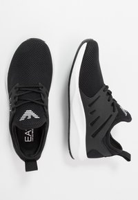 Černé sportovní boty s textilním svrškem z mesh materiálu, texturovanými detaily, zaoblenou špičkou, bílou podrážkou a kontrastním logem na jazyku. Šněrovací design.