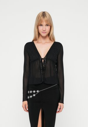 Blouse noire transparente avec un corps en dentelle texturée, décolleté en V profond et nœud à l'avant. Manches longues avec une finition lisse ; portée sur une jupe noire ajustée.