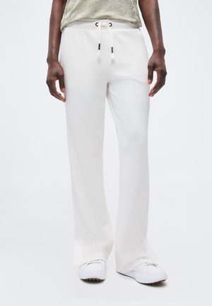 Homme portant un pantalon large blanc avec taille à cordon de serrage et des baskets blanches, debout devant un fond clair uni.