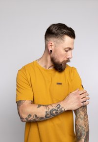 T-shirt di cotone giallo con taschino sul petto, caratterizzata da un piccolo logo ricamato. Il modello ha tatuaggi su entrambe le braccia e un'acconciatura corta.