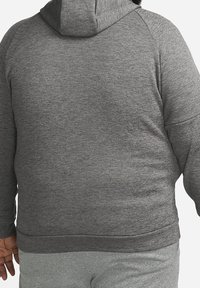 Personne portant un sweat à capuche gris foncé texturé et un pantalon de jogging gris clair assorti, vue de dos sur un fond uni.