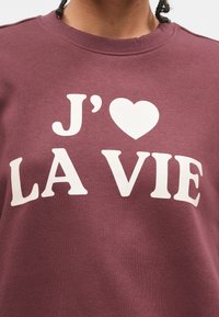 Maroon sweatshirt with white printed text reading "J'❤️LA VIE" in a bold, slightly curved font. Features a classic crew neckline.