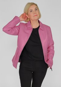 Roze leren jas met een kraag, vooraan met een rits en zijzakken. Gedragen over een zwarte top en gecombineerd met zwarte broek. Glad oppervlak.