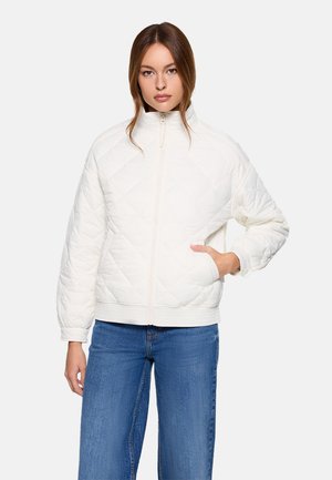 Mujer con chaqueta blanca acolchada con cremallera y manos en los bolsillos, y vaqueros azules, de pie frente a un fondo claro y liso.